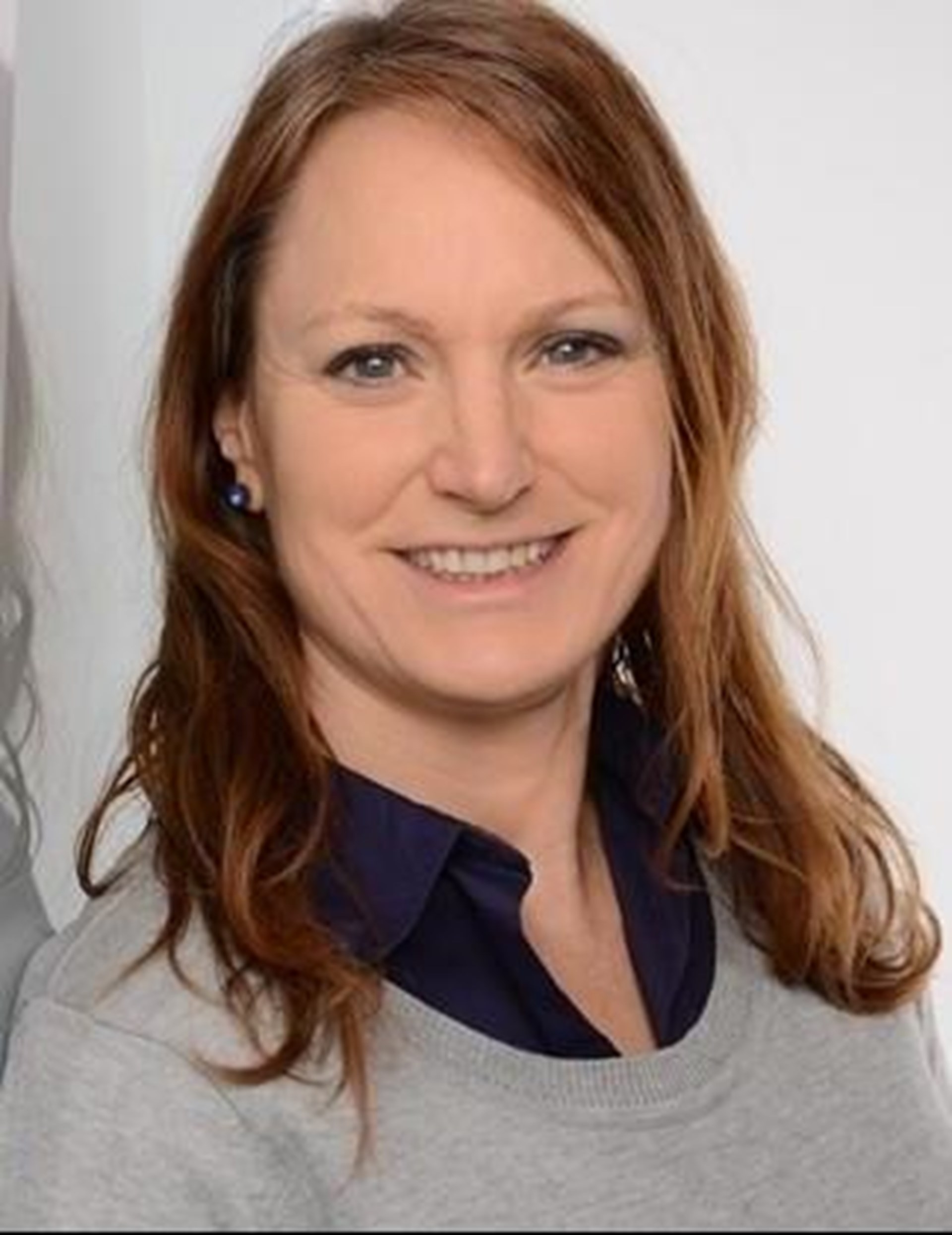 Eine Frau mit schulterlangem, leicht welligem braunem Haar lächelt in die Kamera. Sie trägt einen grauen Pullover über einem dunkelblauen Hemd und steht neben einer spiegelnden Glasfläche.