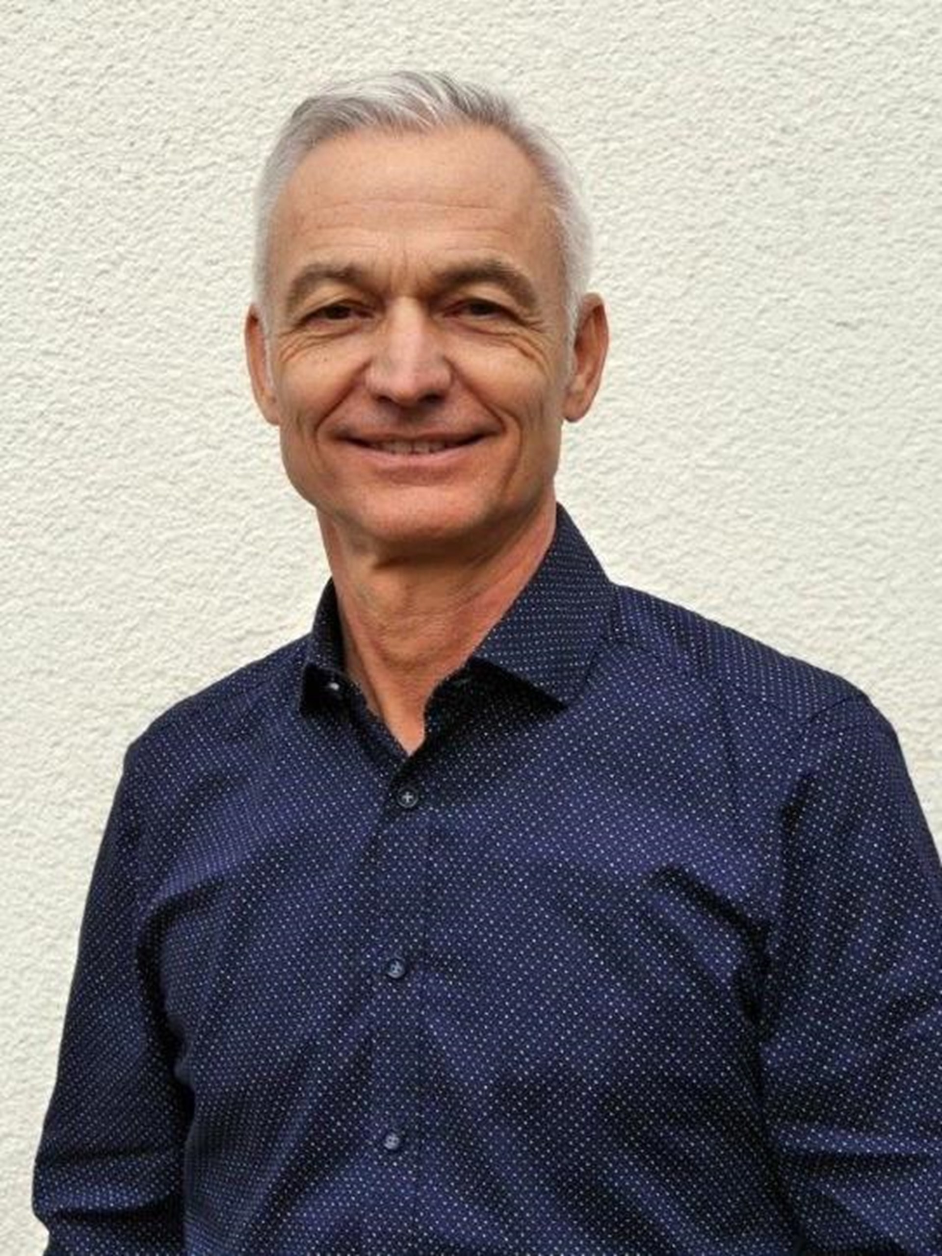 Ein älterer Mann mit grauem Haaren und einem freundlichen Lächeln trägt ein dunkelblaues Hemd mit feinem Muster und steht vor einer weißen Wand.