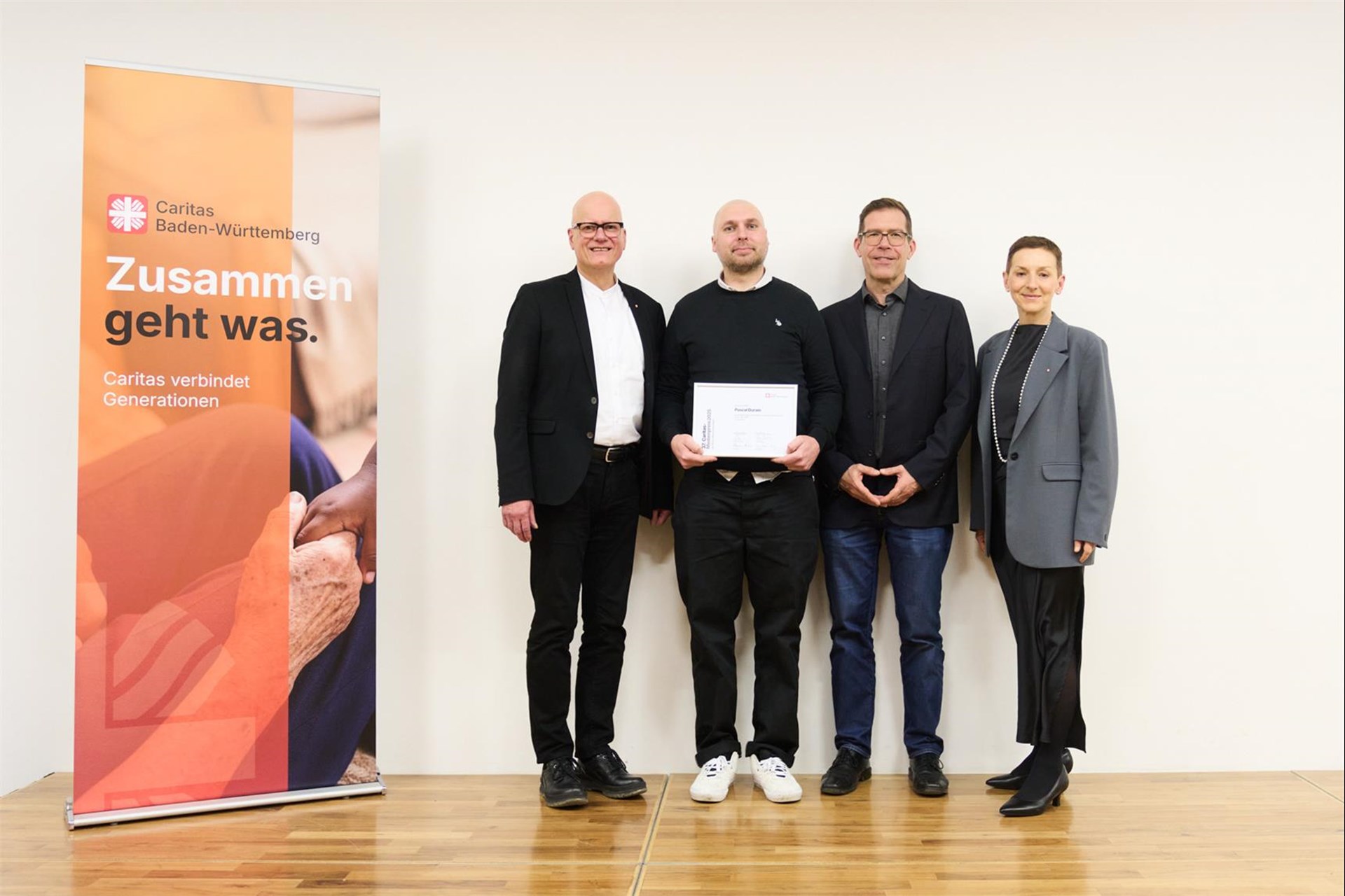 Vier Personen stehen mit einer Urkunde vor einem Caritas Baden-Württemberg vor einem Banner.