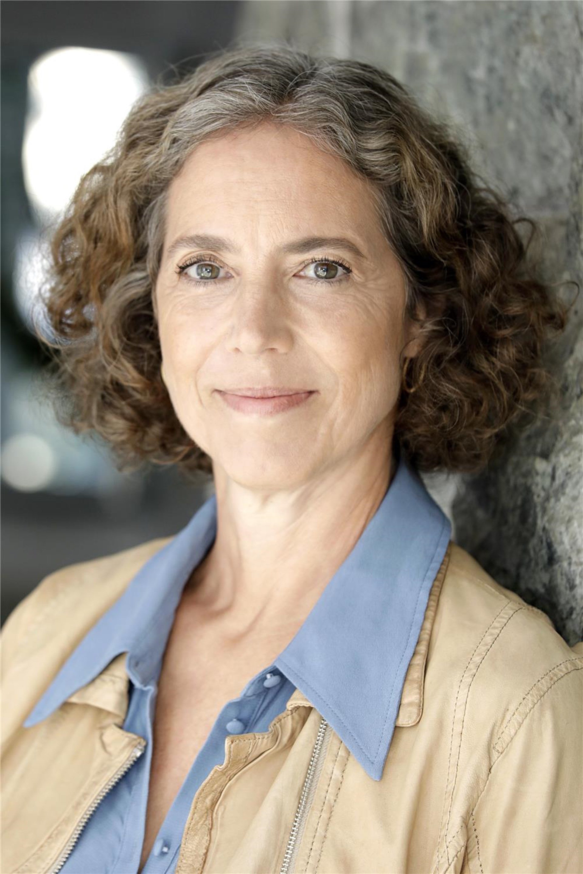  Eine Frau mit lockigem, schulterlangem, graubraunem Haar und einem sanften Lächeln trägt eine beige Jacke über einer hellblauen Bluse und steht vor einer Steinwand.