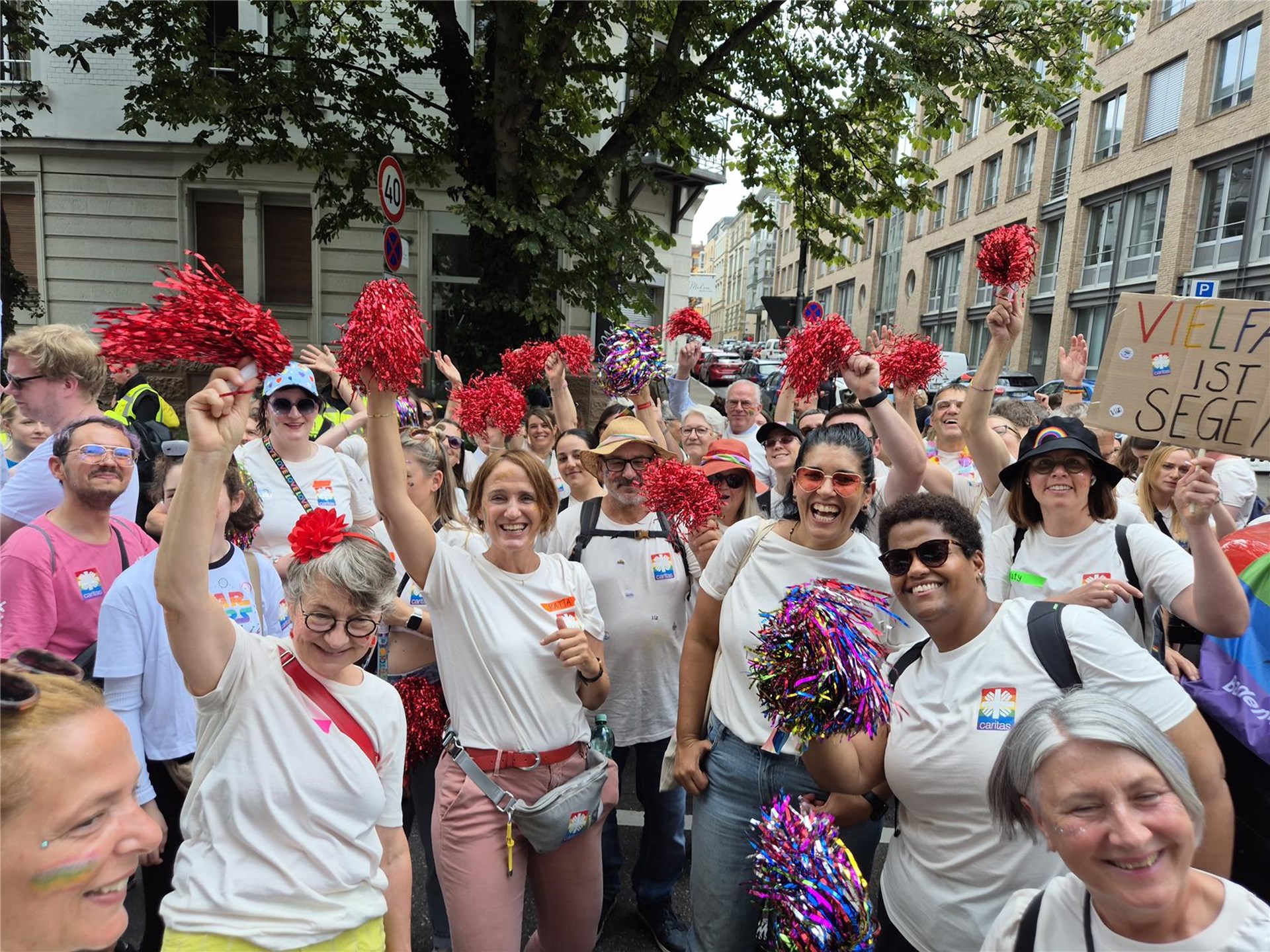 Mehrere Menschen bei einer Pride-Parade winken und lachen in die Kamera. Im Vordergrund eine Person mit Regenbogenflagge und Caritas-T-Shirt.