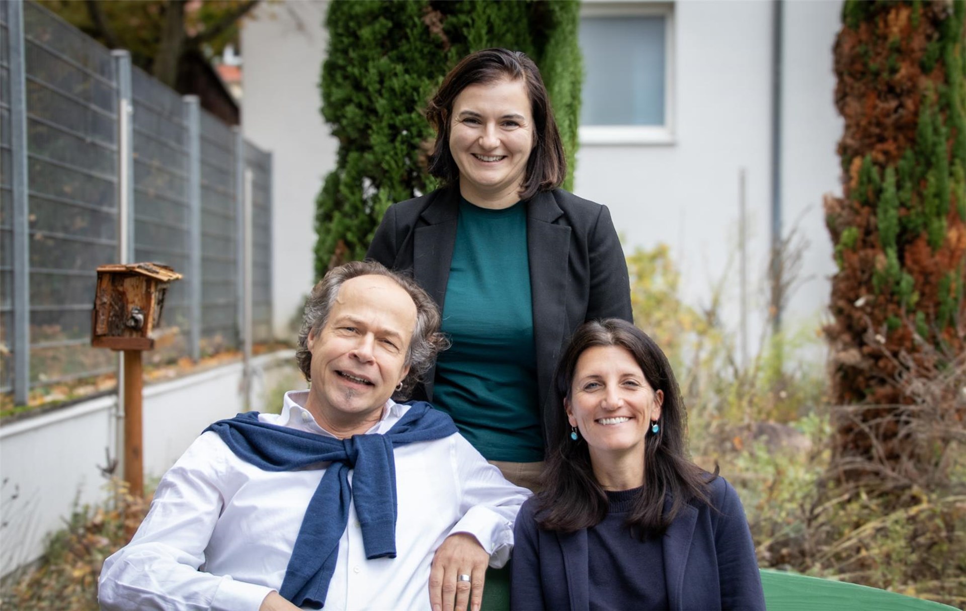 Drei Erwachsene lächeln draußen vor Büschen und einem Wohnhaus.