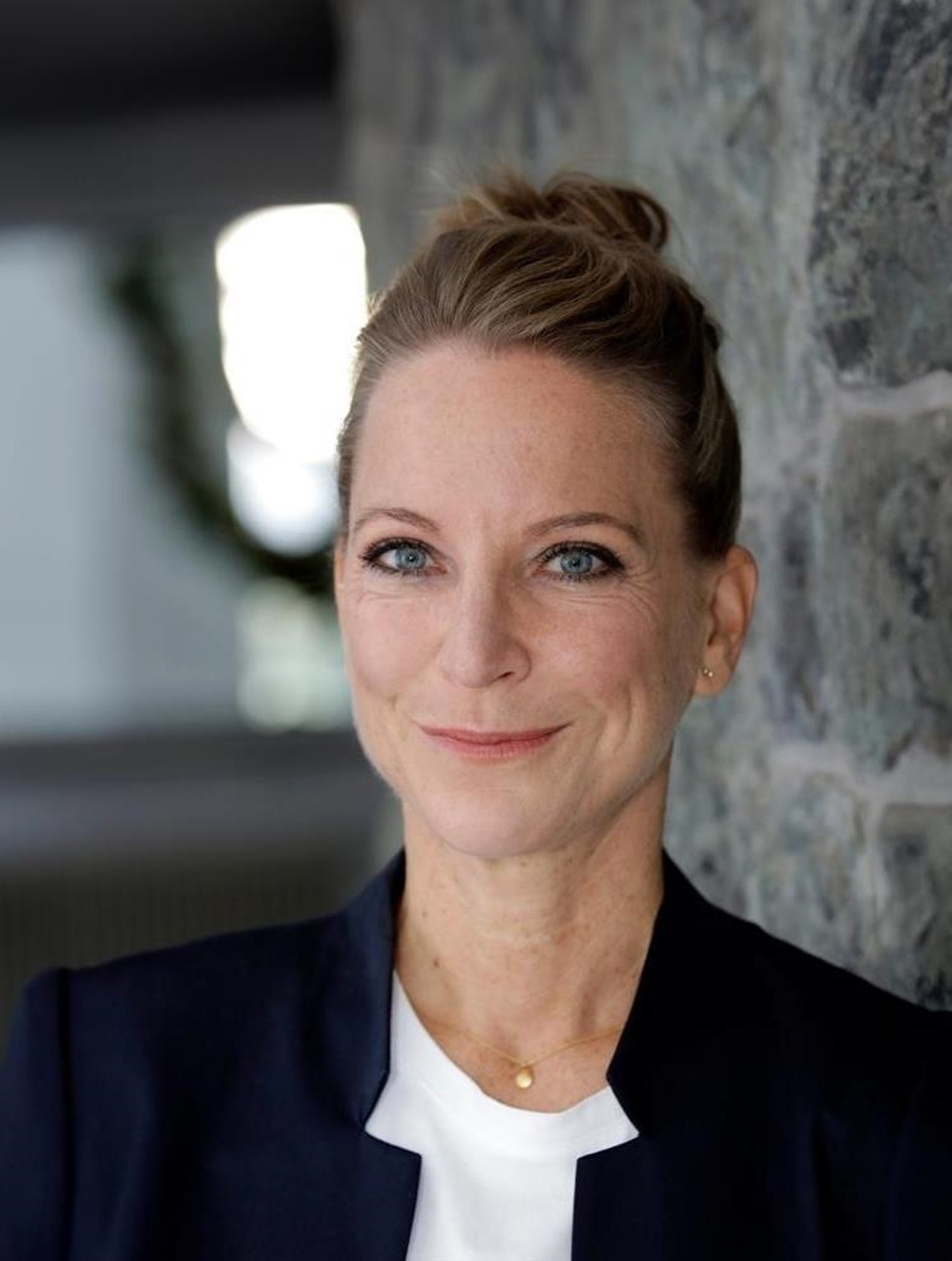 Porträt einer lächelnden Frau mit blonden Haaren, die in einem Dutt gebunden sind. Sie trägt einen dunkelblauen Blazer und ein weißes Oberteil und steht vor einer Steinmauer.