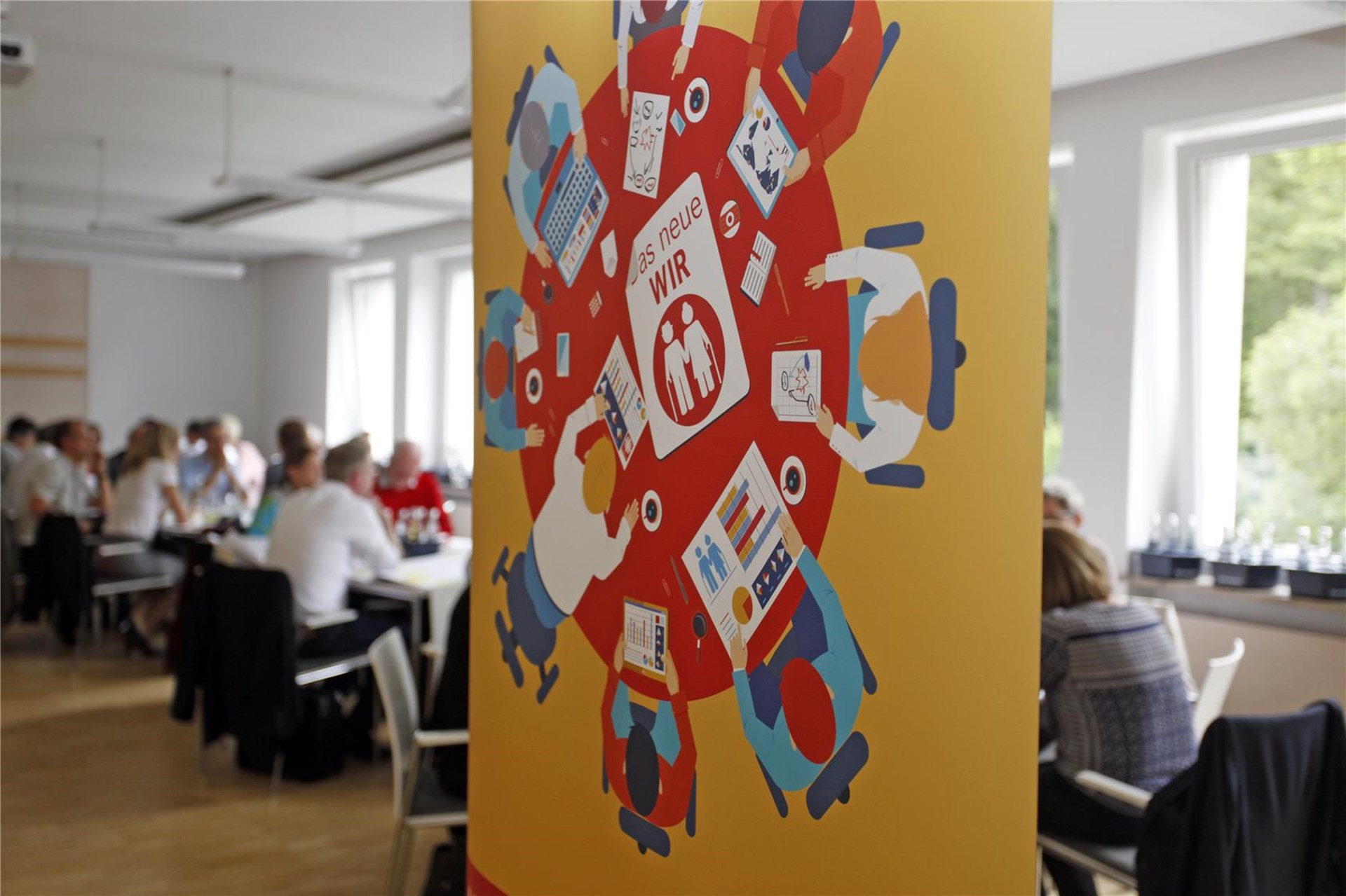 Im Hintergrund: sitzen Menschen um die Arbeitstische herum, Im Vordergrun: Logo des Netzwerkes Alter und Pflege: ein Gemälde bei dem Frauen und Männer um einen runden Tisch  herumsitzen